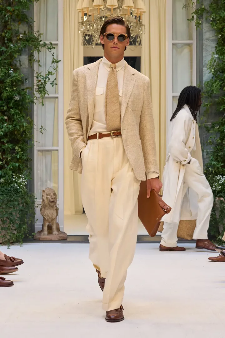 Ralph Lauren İlkbahar 2027 Erkek Giyim koleksiyonundan bir model, bej keten takım elbisesi, krem rengi gömleği, kahverengi tokalı ayakkabıları ve elinde tuttuğu kahverengi deri çantasıyla lüks bir podyumda güneş gözlüklü olarak yürüyor; arka planda yemyeşil sarmaşıklar, klasik bir aslan heykeli ve zarif bir avize görülüyor.