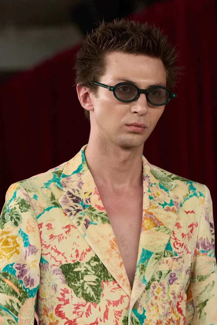 Junya Watanabe'nin 2026 İlkbahar erkek giyim koleksiyonundan, sivri kahverengi saçlı, yeşil çerçeveli oval güneş gözlüklü ve canlı çiçek desenli sarı blazer ceket giymiş bir modelin yakın çekim detay görüntüsü.