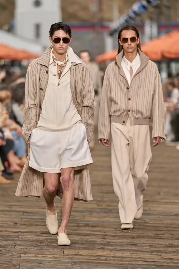 Zegna'nın İlkbahar 2027 erkek giyim koleksiyonundan, podyumda yürüyen iki erkek model, bej, krem ve beyaz tonlarında çizgili ve düz parçalarla oluşturulmuş, güneş gözlükleriyle tamamlanmış zarif ve rahat kıyafetler sergiliyor: sol taraftaki model uzun çizgili bir palto, çizgili polo, beyaz şort ve makosen giyerken, sağdaki model çizgili bomber ceket ve bol bej pantolonla doğal bir ilkbahar stili sunuyor.