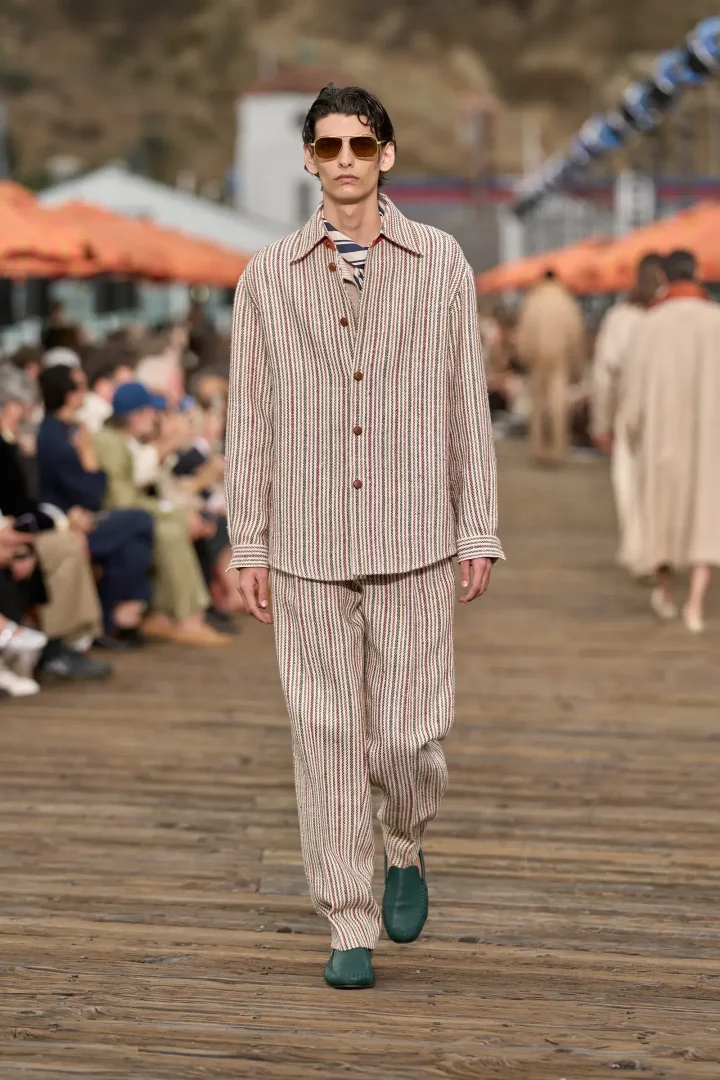 Zegna İlkbahar 2027 erkek giyim koleksiyonundan bir model, açık renk zemin üzerine kırmızı ve yeşil çizgili, bol kesim bir gömlek ve pantolondan oluşan takımı, kahverengi camlı pilot güneş gözlükleri ve turkuaz yeşili makosenleriyle ahşap bir podyumda yürüyor.