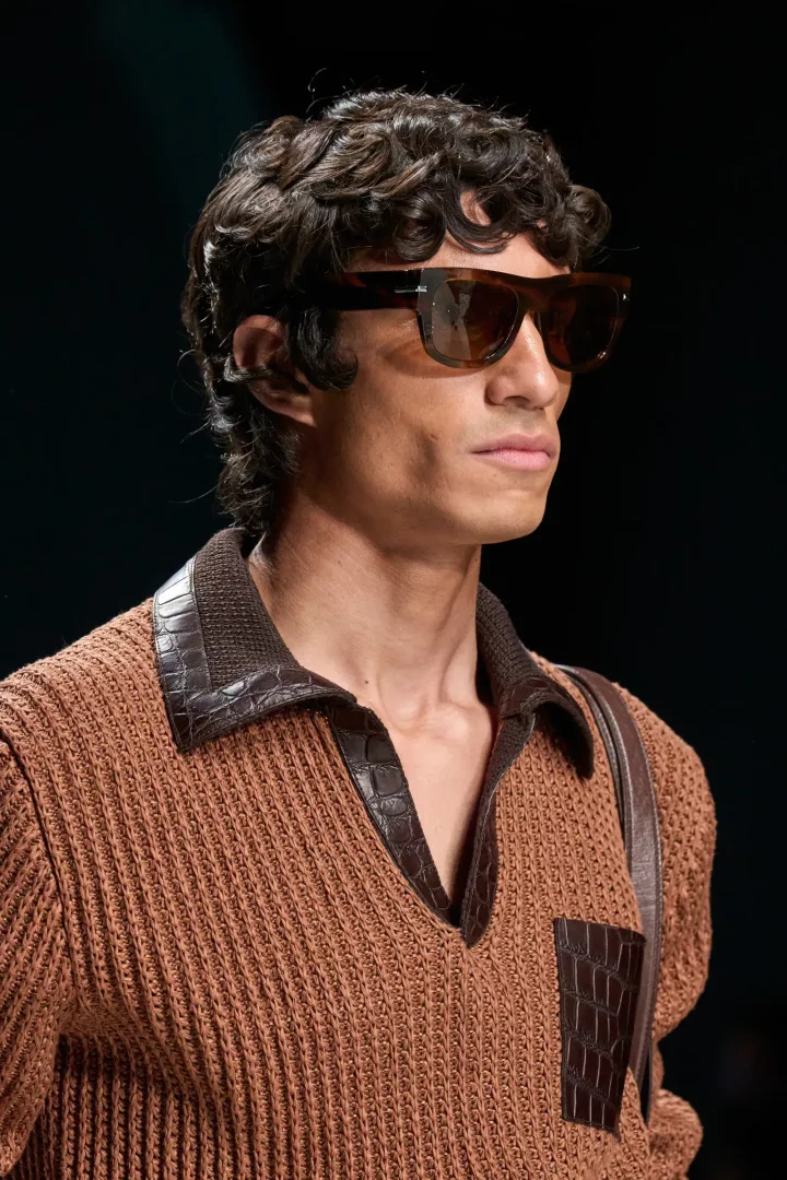 Bir modelin koyu kahverengi, timsah desenli yaka detayına sahip bakır rengi triko polo tişörtü ve şık güneş gözlükleriyle görüldüğü bu yakın plan çekim, Dolce and Gabbana İlkbahar 2027 erkek giyim koleksiyonunun zengin detaylarını sergiliyor.