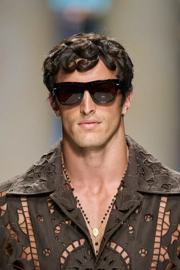 Dolce and Gabbana İlkbahar 2027 erkek giyim koleksiyonundan bir detay: Koyu renk güneş gözlüğü, boynunda altın detaylı siyah boncuklu bir kolye ve yakası açık, lazer kesim desenli kahverengi süet görünümlü bir gömlek giymiş, kıvırcık saçlı bir modelin göğüs hizasından çekilmiş yakın plan portresi.
