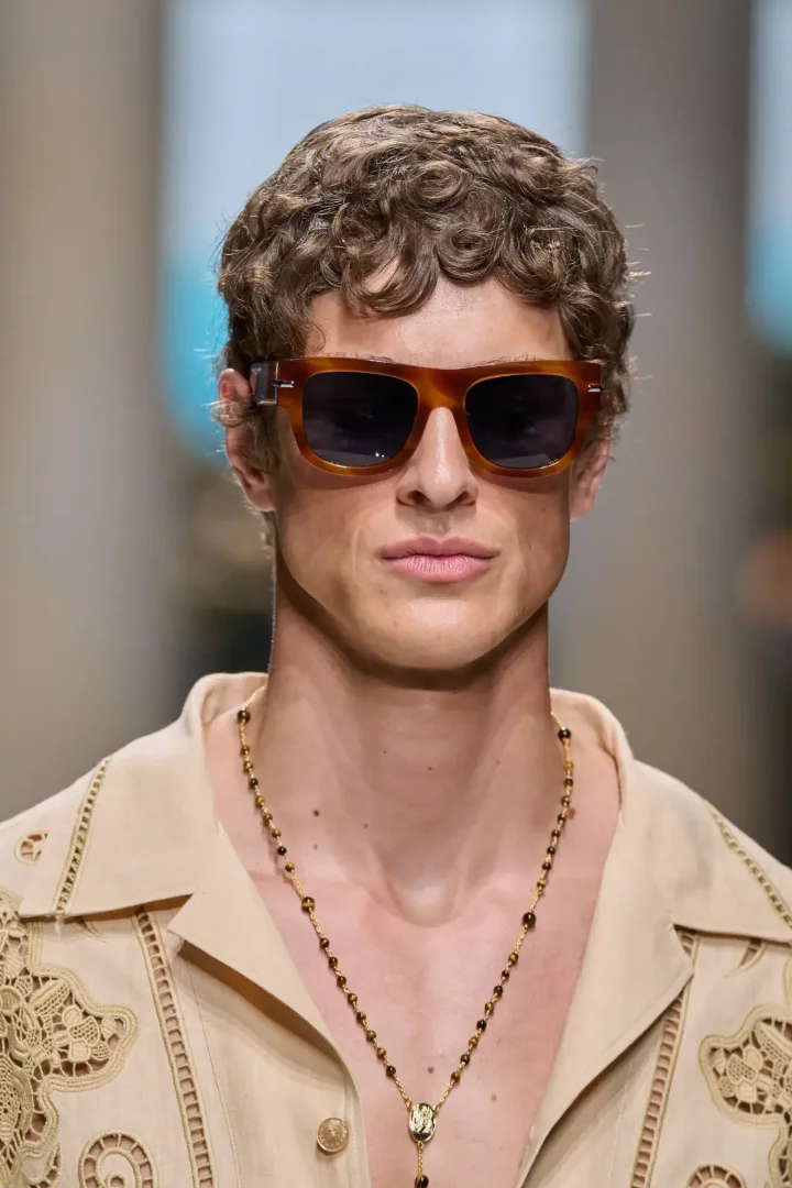 Dolce and Gabbana Bahar 2027 erkek giyim koleksiyonundan bir detayı vurgulayan bu yakın planda, kaplumbağa kabuğu desenli güneş gözlüklü, kıvırcık saçlı bir erkek model, dantel işlemeli bej bir gömlek ve altın madalyonlu kehribar boncuklu uzun bir kolye ile şık ve modern bir görünüm sergiliyor.