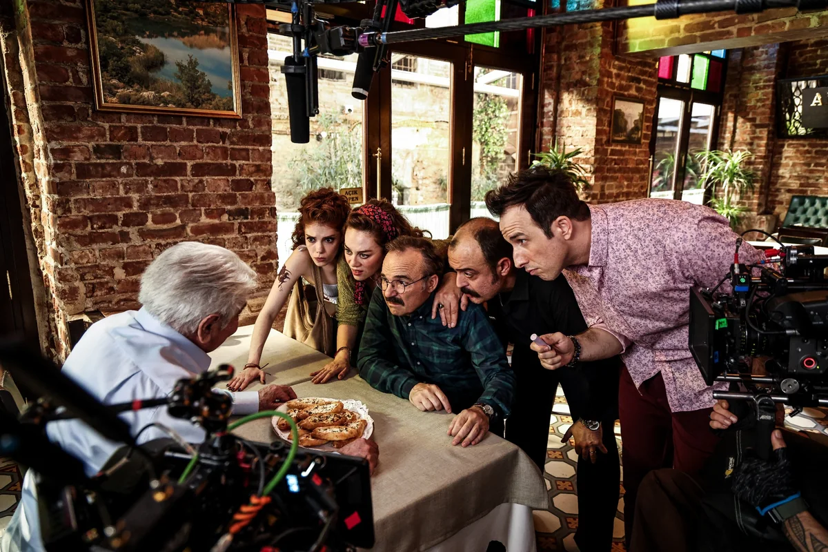 Aile Arasında filminden bir restoran sahnesi set karesinde, oyuncular ve film ekibi, masadaki pide benzeri yiyeceklere veya yaşlı bir adama dikkatle bakarken, ön planda çekim ekipmanları net bir şekilde görülüyor.