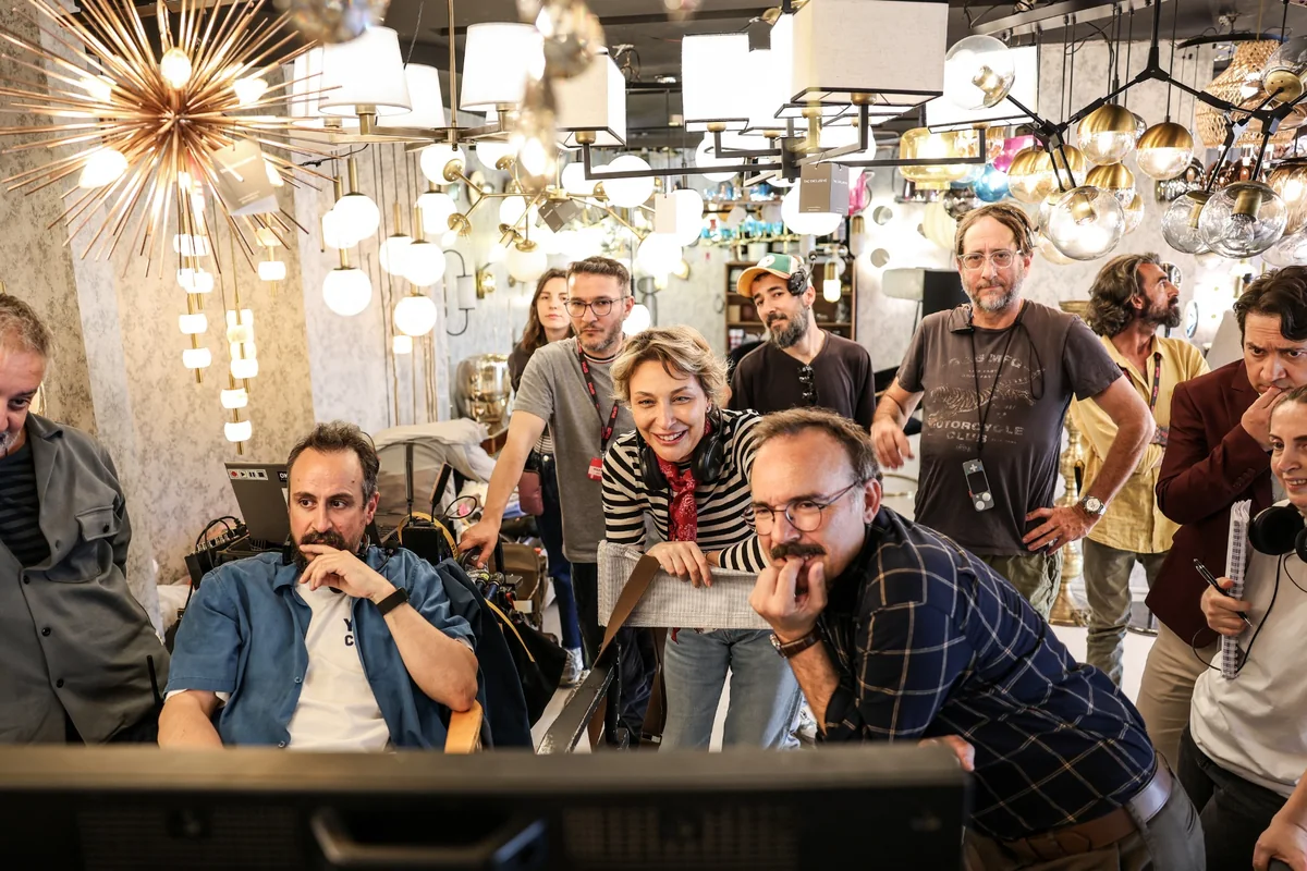 Aile Arasında filminin set arkası çekim ekibi, arka planda birçok dekoratif aydınlatma armatürünün olduğu bir mekanda, bir monitöre odaklanmış, aralarında gülümseyen bir kadın ve sakallı erkeklerin de bulunduğu bir grup olarak sahneyi izlerken.