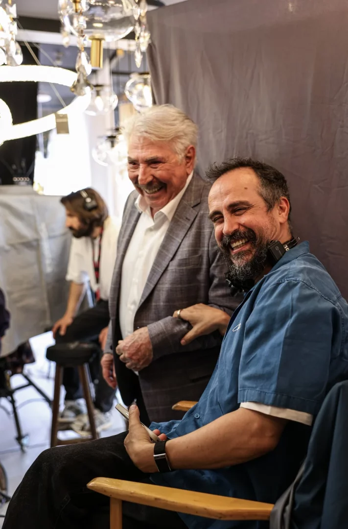 Aile Arasında filminin neşeli set arkası görüntüsünde, beyaz saçlı ve çizgili ceketli bir oyuncu ile kulaklığı boynunda, mavi gömlekli filmin yönetmeni içten kahkahalarla kameraya bakarken görülüyor; oyuncunun yönetmenin koluna girdiği samimi poz, çekim ortamının dostane ve keyifli atmosferini yansıtıyor, arkada ise set ekipmanları ve belirsiz bir çalışan yer alıyor.