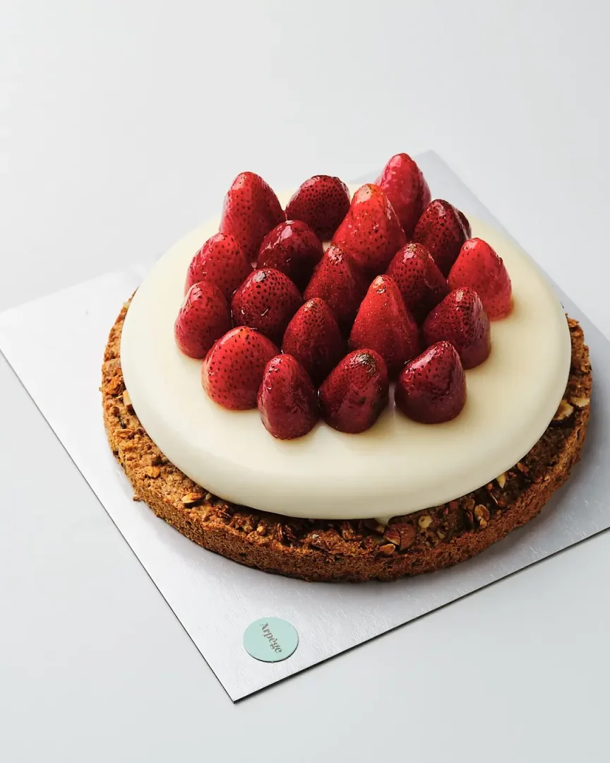 Gümüş renkli bir servis tabağının üzerinde duran, bol ve parlak taze çileklerle kaplı beyaz kremsi bir katmana ve altında fındık parçacıklı kıtır bir tabana sahip, Arpège Patisserie imzalı iştah açıcı çilekli tart.