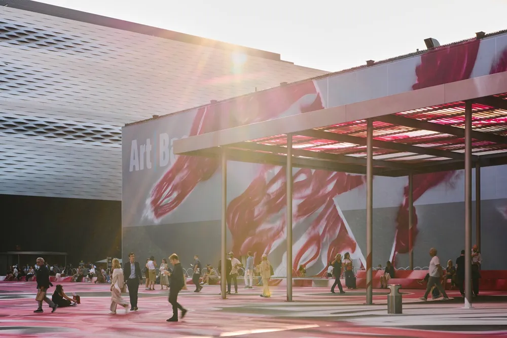 Parlak güneş ışığının vurduğu Art Basel fuar alanında, zemini kırmızı, pembe ve beyaz soyut desenlerle kaplı geniş bir meydanda ziyaretçiler gezinirken, arka planda Art Basel yazılı dev bir pankart ve çağdaş binalar dikkat çekiyor.