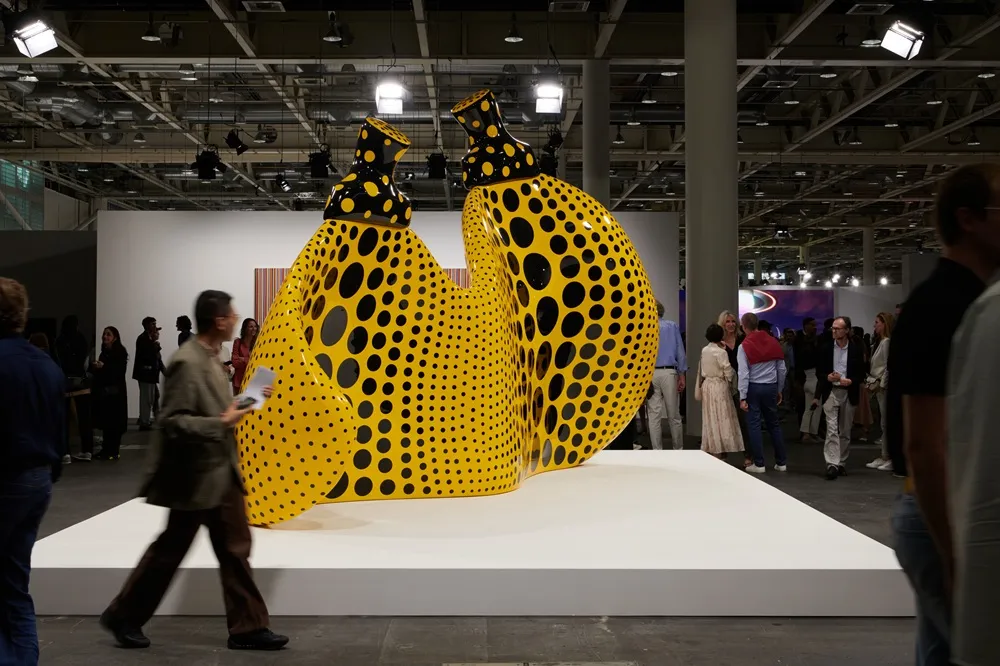 Art Basel etkinliğinde, geniş beyaz bir platform üzerinde sergilenen, organik formlu, parlak sarı ve siyah puantiyelerle kaplı iki devasa heykel ile etraflarındaki kalabalık sanatseverleri gösteren genel bir görünüm.