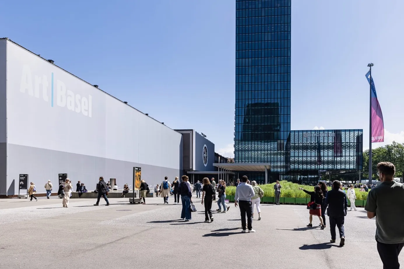 Güneşli bir havada, Art Basel fuarının hareketli girişinde, üzerinde Art Basel yazan geniş beyaz sergi binası ve yansımalı bir cam gökdelen önünde birçok kişinin fuar alanına doğru ilerlediği, canlı bir etkinlik ortamı sergileniyor.