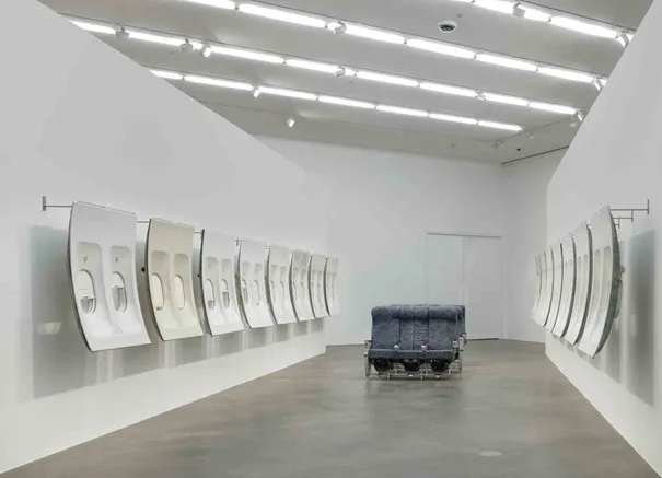 Beyaz duvarlı modern bir sanat galerisinde, Art Basel kapsamında sergilenen, her iki duvarda karşılıklı sıralanmış, uçak pencere bölmelerini andıran beyaz paneller ve ortada tekerlekli bir platform üzerinde duran koyu renk, dikdörtgen, endüstriyel görünümlü bir heykelden oluşan çağdaş bir enstalasyon.