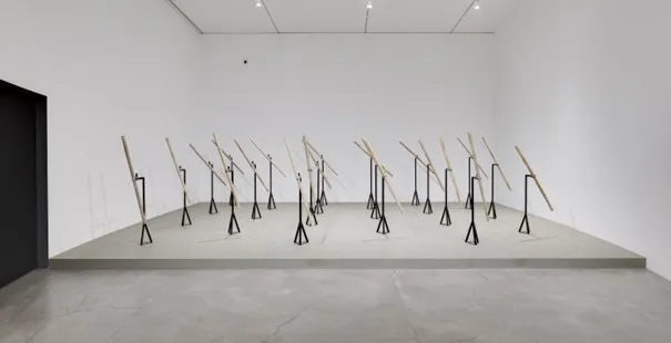 Art Basel'in görülmesi gerekenler listesinden bir parça olarak, beyaz duvarlı minimalist bir galeride gri yükseltilmiş bir platform üzerinde sıralanmış, siyah üçgen metal ayaklar üzerinde eğimli duran açık renkli ahşap çubuklardan oluşan soyut bir enstalasyon sergileniyor.