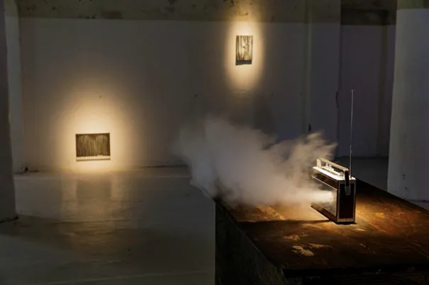 Bu enstalasyon, Art Basel'in görülmesi gerekenler listesinde öne çıkanlardan biri olarak, loş ışıklı bir sergi alanında ahşap bir masa üzerinde yoğun duman çıkaran antenli eski model bir radyo ve duvarda spot ışıklarıyla aydınlatılmış iki eseri gösteriyor.