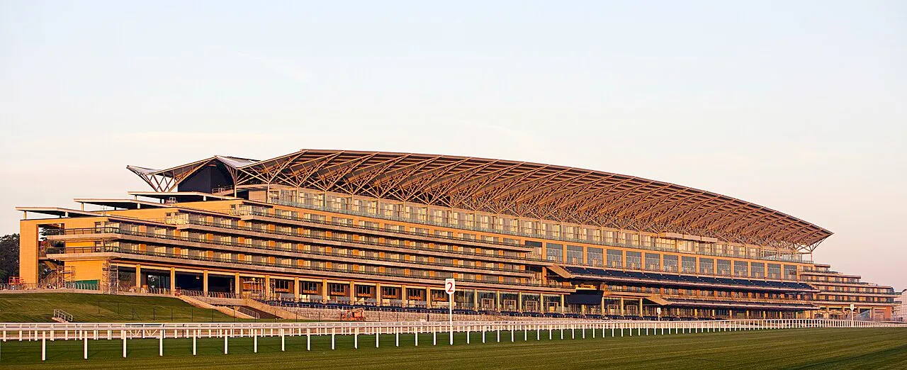 Ascot Hipodromu'nun geniş yeşil çim pistinin hemen arkasında yükselen, çağdaş mimariye sahip, metal iskeletli kavisli çatısıyla dikkat çeken çok katlı, modern tribün binası gün batımı ışıkları altında parlıyor.