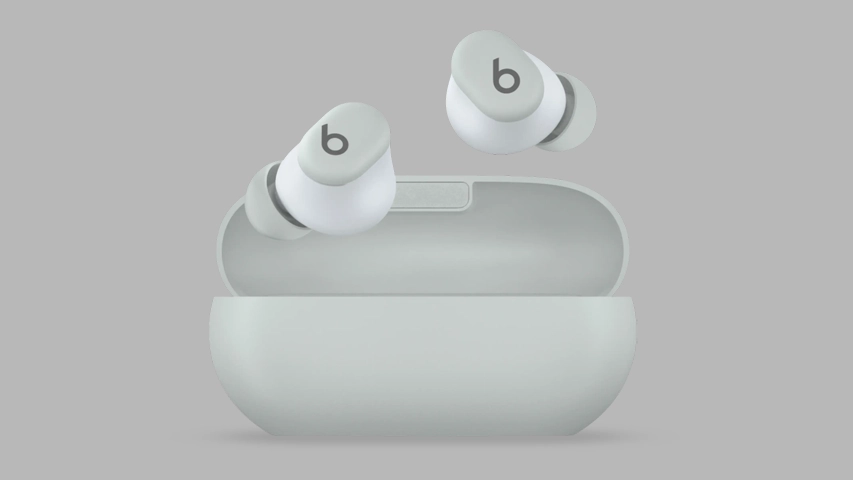 İki adet açık gri renkte Beats Studio Buds kablosuz kulaklık, aynı renk şarj kutusunun açık kapağının üzerinde havada asılı duruyor; kulaklıkların dış yüzeyinde Beats logosu olan küçük b harfi belirgin şekilde görülüyor ve genel tasarım pürüzsüz hatlara sahip.