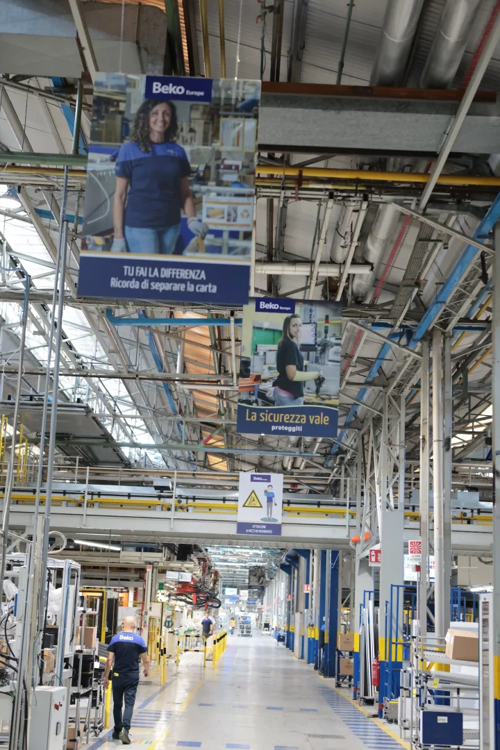 Beko Europe markalı iş kıyafetleri giyen çalışanların yürüdüğü, modern makineler ve İtalyanca güvenlik ile sürdürülebilirlik mesajları içeren afişlerle dolu geniş bir fabrika üretim hattı, Arçelik'in Avrupa'daki üretim ayak izi ve küresel rekabetteki stratejik gücünü temsil eden Cassinetta İşletmeleri'nden bir kesit sunuyor.