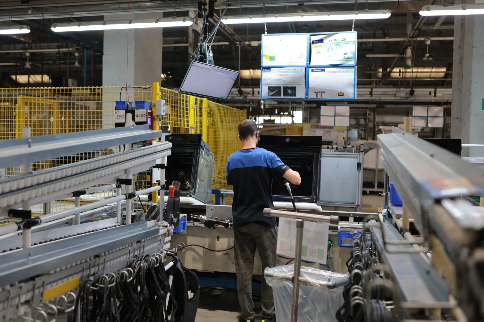 Beko Europe Cassinetta fabrikasında, montaj hattında bir fırının iç aksamlarını özenle birleştiren mavi-siyah tişörtlü bir çalışan, ankastre teknolojilerindeki üretim ayak izini gösteriyor; etrafında konveyör bantlar ve dijital ekranlarda enerji sınıfı bilgileri görülüyor.