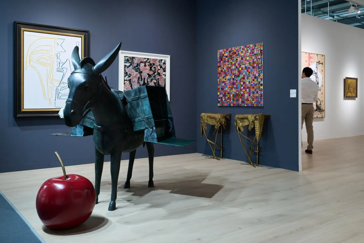Koyu mavi duvarlı bir sanat galerisinde, eyer çantalı bronz bir eşek heykeli ve yanında yerde duran parlak kırmızı, büyük bir kiraz heykeli ön planda; arka planda ise soyut tablolar, duvara monte timsah figürlü sehpalar ve galeride dolaşan bir ziyaretçi yer alıyor.