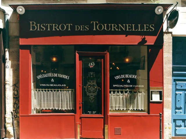 Kırmızı cepheli, ön cephesinde BISTROT des TOURNELLES tabelası bulunan klasik Fransız restoranının vitrinlerinde Beaujolais şarapları ve özel et spesiyaliteleri sunulduğunu belirten yazılar, işlemeli cam kapısı ve dantel perdelerle sıcak bir karşılama hissi uyandıran zarif bir dış görünüm.