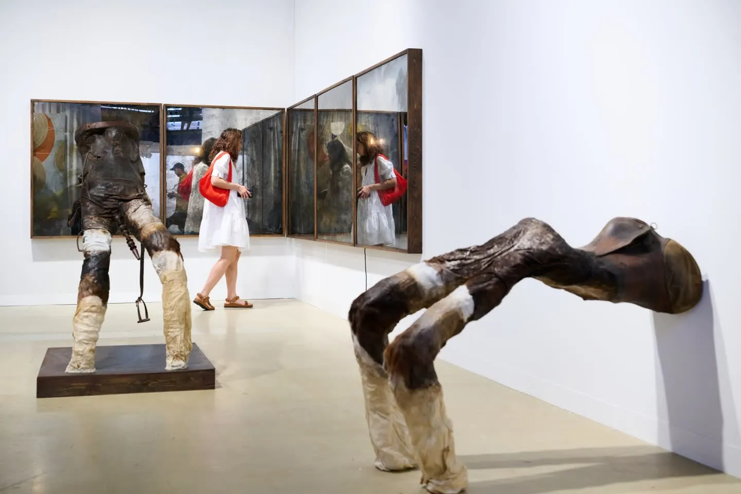 Beyaz elbiseli ve kırmızı omuz çantalı bir ziyaretçi, sanat sergisi alanında yansıtıcı yüzeylere sahip üç büyük tabloya bakarken, ön planda kahverengi ve krem renklerinde, dokulu, biri kaide üzerinde ayakta duran diğeri duvara yaslanmış at gövdesi ve bacaklarını andıran iki soyut heykel görücüye çıkıyor.