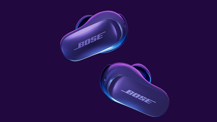 Koyu mor-mavi tonlarındaki iki adet ikinci nesil Bose QuietComfort Ultra kulaklık, üzerinde parlak beyaz BOSE logosu ve kenarlarında ince parlak mavi hatlar ile modern tasarımlarını vurgulayarak koyu mor bir arka plan üzerinde havada süzülür gibi görünüyor.