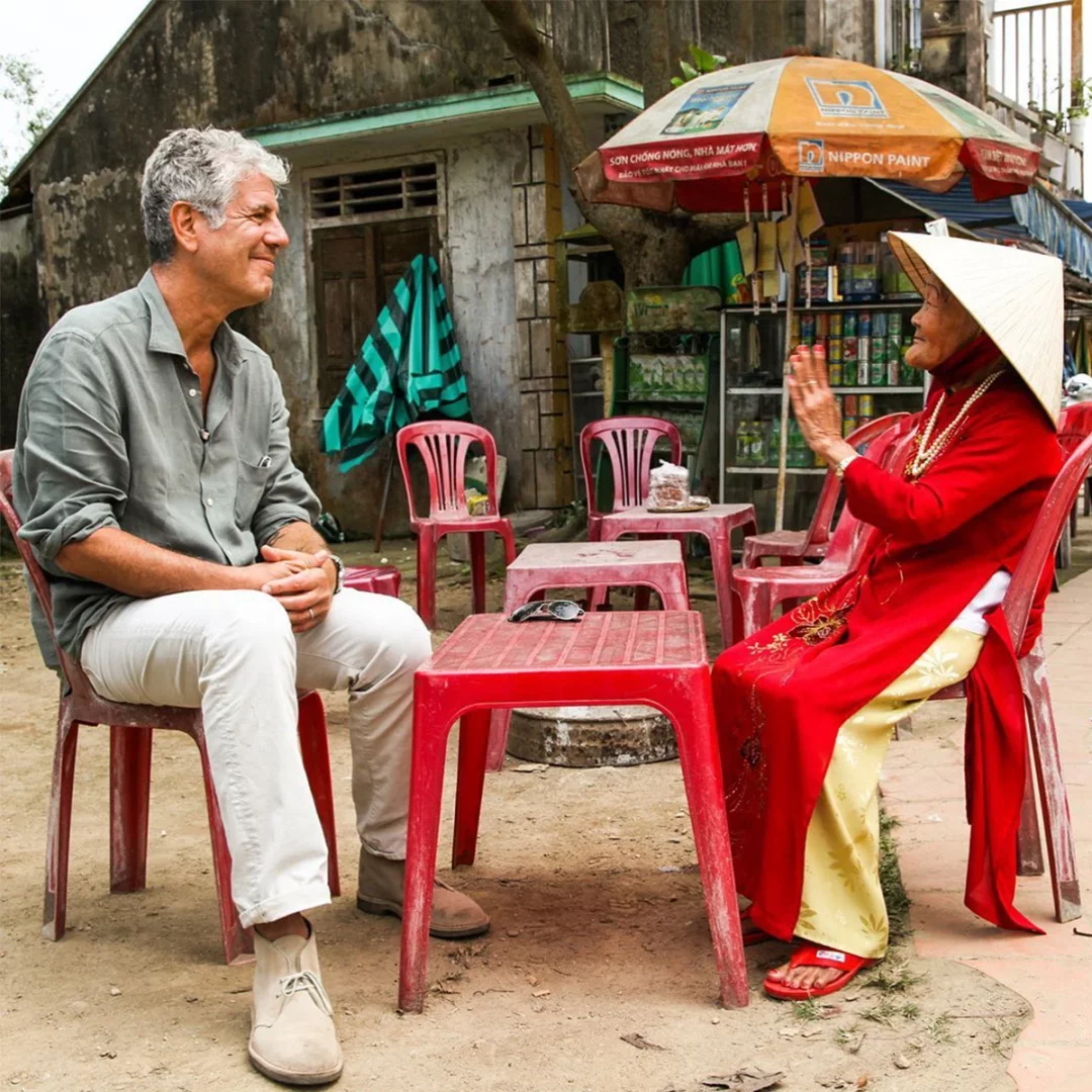 Gri saçlı Anthony Bourdain, krem rengi pantolonu ve açık renk gömleğiyle sol tarafta kırmızı bir sandalyede oturmuş, karşısındaki geleneksel kırmızı áo dài giymiş, hasır konik şapkalı yaşlı Asyalı kadını ilgiyle dinlerken hafifçe gülümsüyor; kadın, ellerini yukarı kaldırarak neşeyle konuşuyor ve ikili, rustik bir binanın önünde, açık havada küçük kırmızı masalarda oturuyor.
