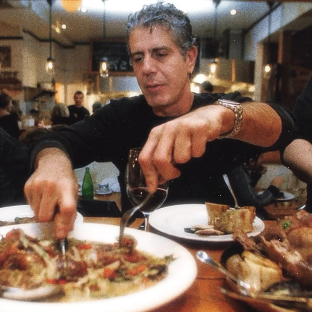 Anthony Bourdain, bir restoranda, elinde çatal ve bıçakla önündeki bol malzemeli yemeği ustalıkla kesip büyük bir iştahla yemeye hazırlanırken, masadaki kırmızı şarap kadehi ve çevresindeki rahat atmosfer, onun eşsiz yemek keşiflerini yansıtıyor.