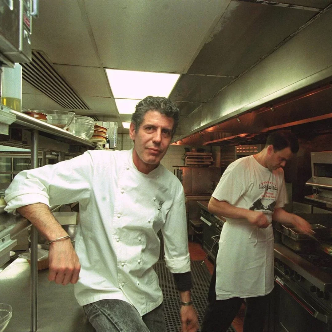 Anthony Bourdain, beyaz şef ceketini giymiş ve ciddi bir ifadeyle doğrudan kameraya bakarken, yoğun bir mutfak ortamında metal bir tezgaha yaslanmış, arka planda bir başka aşçı da yemekle meşgul.