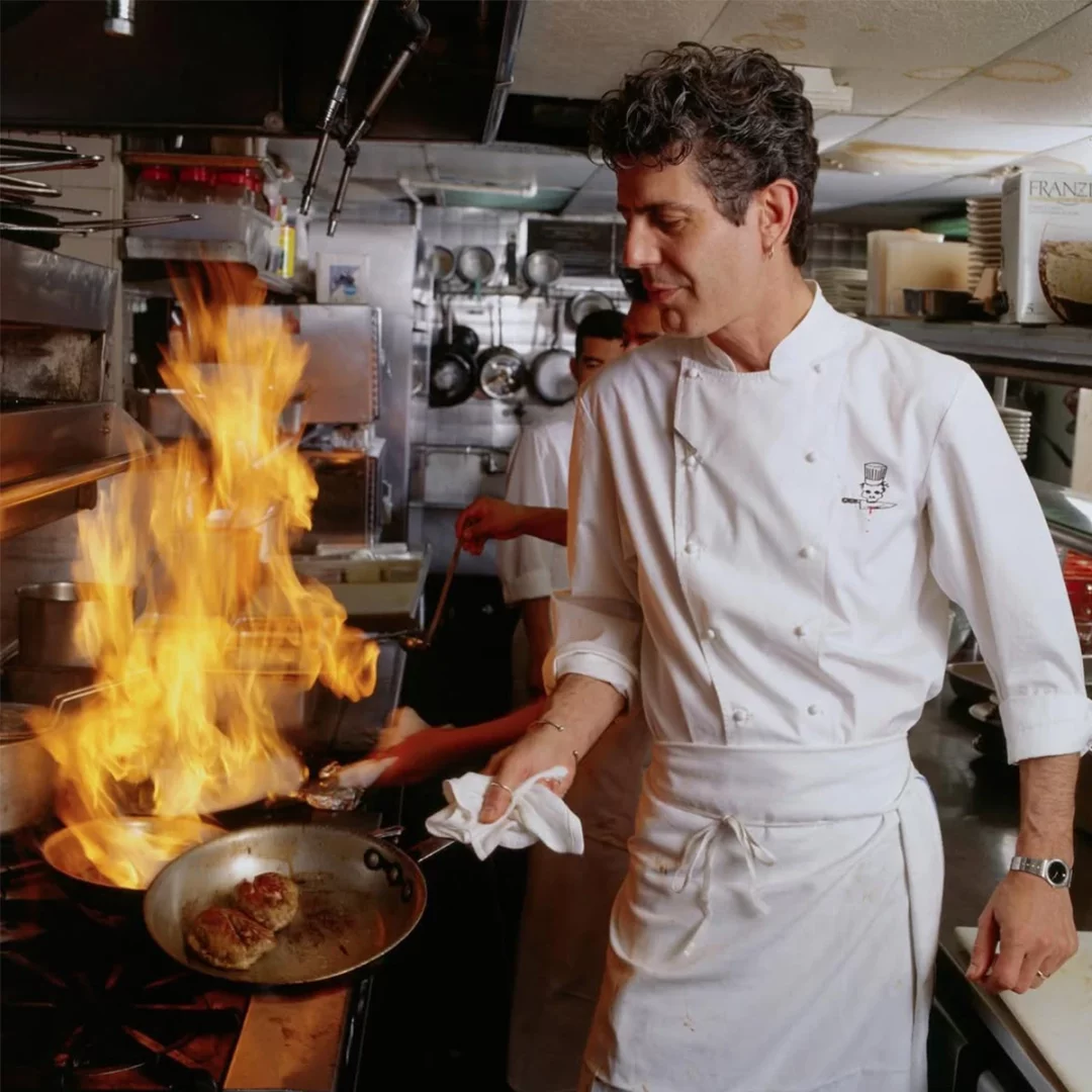 Anthony Bourdain, profesyonel bir mutfakta, tencereden yükselen büyük, parlak alevlerin eşliğinde yoğun bir şekilde yemek pişirirken, karakteristik dalgalı saçları ve şef üniformasıyla elinde beyaz bir bez tutarak yaptığı işe odaklanmış bir şekilde görünüyor.