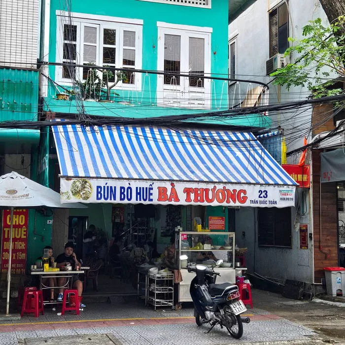 Bún Bò Huế Bà Thương adlı Vietnam restoranının turkuaz renkli binasının önünde, mavi beyaz çizgili tentenin altında masalarda oturup yemek yiyen insanları, yiyecek standını ve parkedilmiş bir motosikleti çevreleyen yoğun elektrik kablolarıyla dolu hareketli bir sokak görünümü.