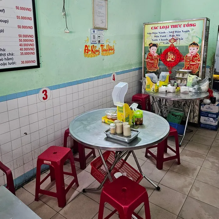Aydınlık yeşil duvarları, beyaz fayans kaplamaları ve kırmızı plastik tabureleriyle sade bir restorana benzeyen bir mekanda, üzerinde baharatlık tepsisi bulunan yuvarlak metal bir masa ile sağda içecek dolabı ve geleneksel süslemeler yer alıyor; bu görüntü Vietnam mutfağını sunan bir Kim Chau gibi bir yerin sıcak ve otantik atmosferini yansıtıyor.