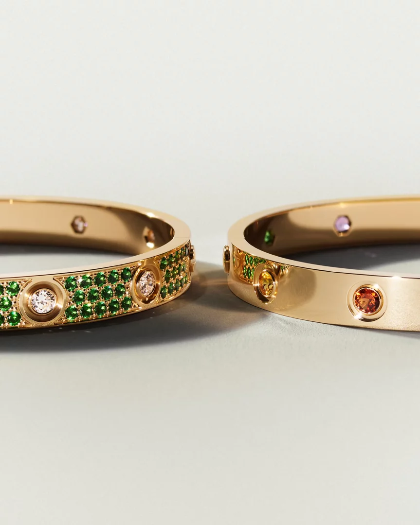 Yakından çekilmiş bir çift Cartier Love koleksiyonu bileziği; soldaki yeşil zümrütler ve pırlantalarla süslenmişken, sağdaki altın renkli bilezikte sarı, turuncu ve mora çalan değerli taşlar ışıltılı bir şekilde öne çıkıyor.
