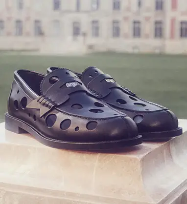 Açık renk bir kaide üzerinde, belirgin deliklere ve yan kısmında şimşek desenli metalik bir detaya sahip, Christian Louboutin'in sonbahar-kış loafer koleksiyonundan modern ve şık siyah ayakkabılar görülüyor.