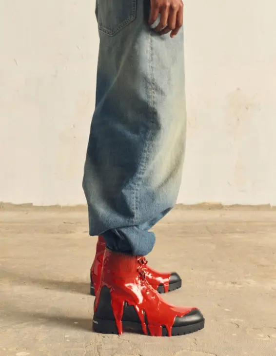 Geniş paçalı açık renk kot pantolon giyen bir kişinin ayaklarında, Christian Louboutin Erkek Sonbahar-Kış koleksiyonundan, siyah tabanlı ve tüm yüzeyine erimiş gibi duran parlak kırmızı malzeme damlalarıyla kaplanmış Molten Trapman botlar dikkat çekiyor