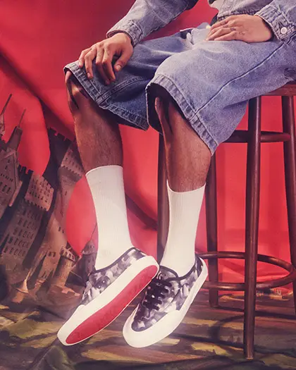 Geniş denim şortlar ve beyaz çorap giyen bir erkeğin, kırmızı arka plan ve kale motifiyle süslenmiş bir dekor önünde, Christian Louboutin'in Sonbahar-Kış koleksiyonundan kamuflaj desenli, ikonik kırmızı tabanlı Tactical spor ayakkabılarını gösterdiği bir fotoğraf.