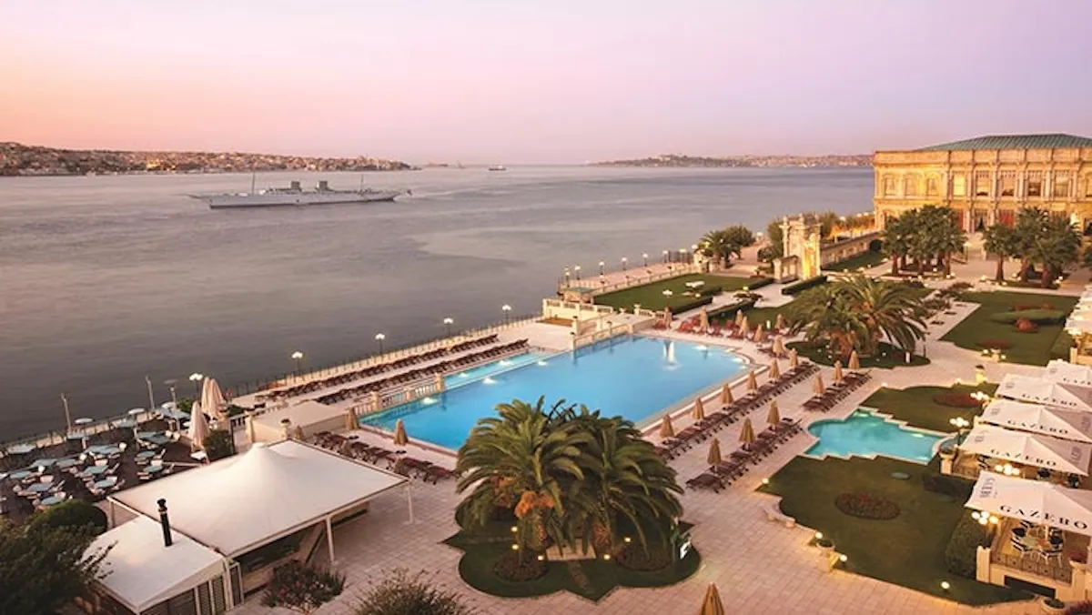 İstanbul Boğazı kıyısında konumlanan Çırağan Sarayı Kempinski'nin genel görünümünde, şezlonglarla dolu ferah açık yüzme havuzu, palmiyelerle süslü bahçeler ve beyaz kameriyelerle çevrili alanlar, günbatımı ışıkları altında Boğaz'da ilerleyen bir gemi ve karşı kıyıdaki İstanbul silüetiyle birleşiyor.