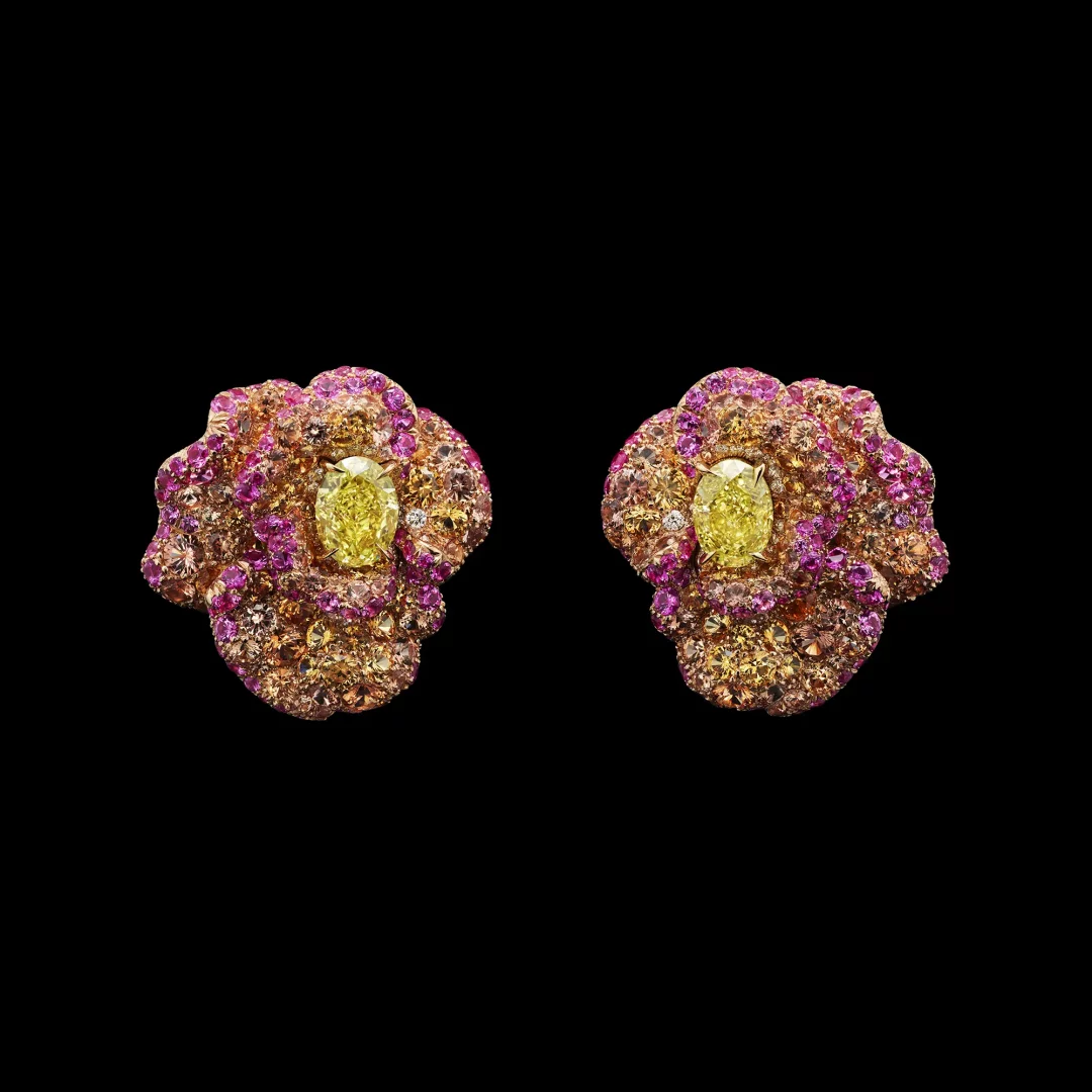 Siyah bir zeminde sergilenen, ortasında parlak sarı oval bir pırlanta bulunan ve pembe, mor, turuncu tonlardaki değerli taşlarla zenginleştirilmiş, çiçeksi motiflere sahip, Dior'un yeni Mücevher Koleksiyonu'ndan göz alıcı bir çift küpe.