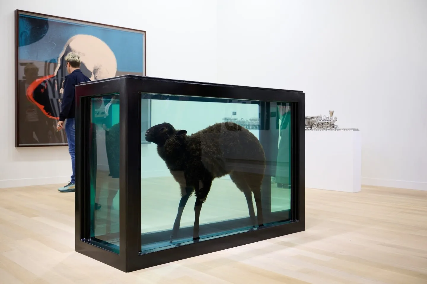 Sanat galerisinin açık renk ahşap zemini üzerinde, siyah çerçeveli cam bir tank içinde yeşilimsi berrak bir sıvıya yerleştirilmiş siyah tüylü bir koyun sanat eseri sergileniyor; sol arkada büyük bir soyut tabloya bakan bir ziyaretçi ve sağda metalik bir heykel ile modern bir sergi atmosferi.