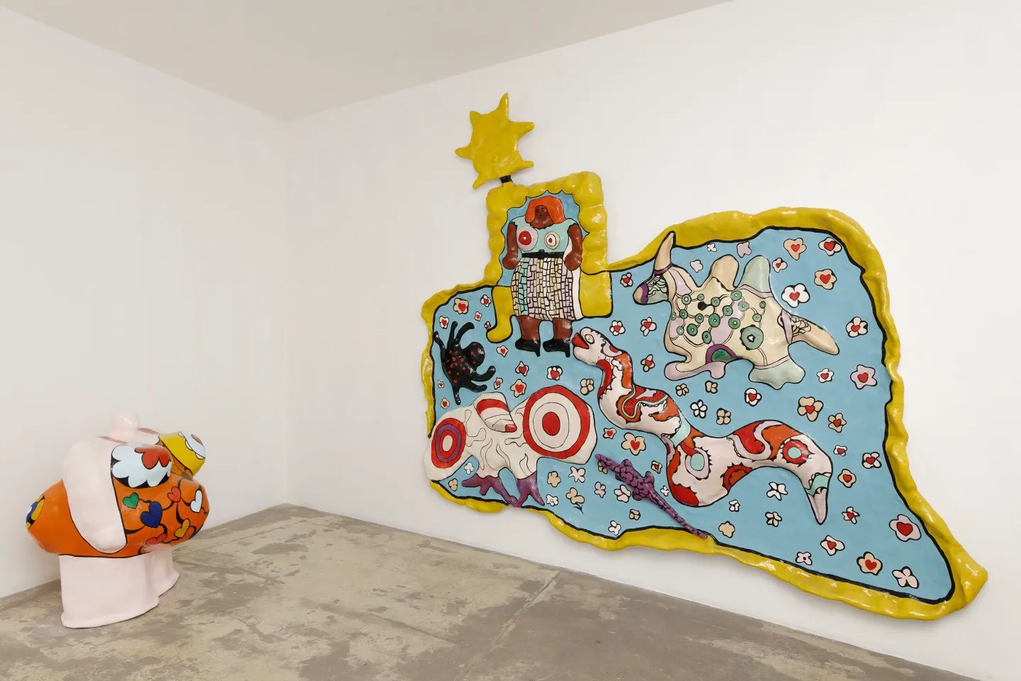 Beyaz duvarlı ve gri zeminli bir sanat galerisi köşesinde, sarı çerçeveli, açık mavi zemin üzerine kırmızı, beyaz, siyah ve yeşil tonlarda yılan, hedef, örümcek gibi çeşitli stilize figürler ile renkli noktaların işlendiği büyük, canlı bir tekstil sanat eseri sergileniyor; eserin üzerinde parlak sarı, ışık hüzmeli bir şekil dikkat çekiyor.