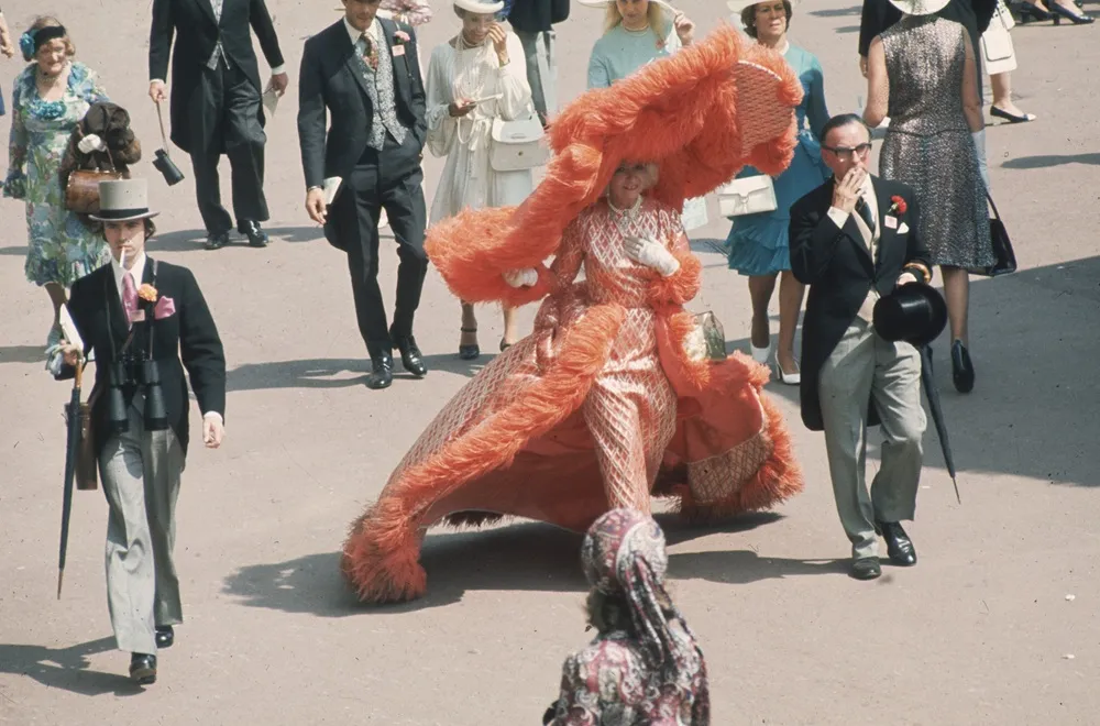 1969 Royal Ascot'ta, Gertrude Shilling'in üzerine turuncu tüylerle bezenmiş, oldukça geniş ve gösterişli bir şapka ile uyumlu, dokulu bir elbise giyerek kalabalık arasında parladığı bu tarihi fotoğrafta, çevresindeki şapkalı, redingotlu ve dürbünlü resmi giyimli kişiler de dikkat çekiyor.