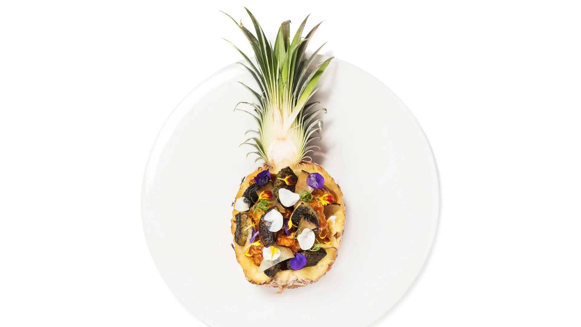 Beyaz bir tabakta, içi çeşitli malzemelerle doldurulmuş, zarifçe hazırlanmış yarım bir ananas yemeği; içerisinde koyu renkli, beyaz parçacıklar, mor ve sarı yenilebilir çiçekler ile yeşil otlarla süslenmiş, egzotik ve modern bir sunum sergileniyor.