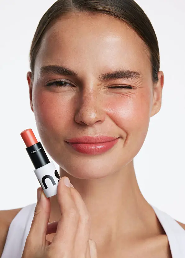 Yanakları ve dudakları doğal pembe tonlarında makyajlı, neşeyle göz kırpan ve gülümseyen genç bir kadın, INO-Beauty için sol elinde Catch Bloom markalı, güzellik rutinine pratik bir dokunuş katacak mercan rengi bir allık/dudak balmı çubuğu tutuyor.