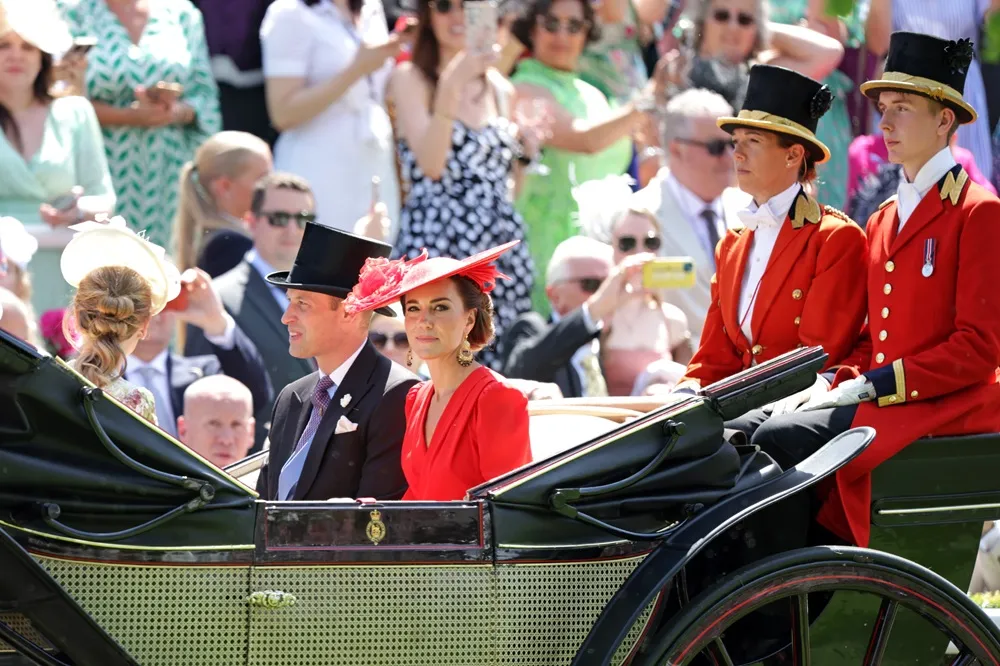 İngiliz yüksek sosyetesinin gözde etkinliği Royal Ascot'ta, Prens William ve kırmızı elbisesiyle Prenses Catherine'in şık bir at arabasında ilerlerken, arkalarında kırmızı üniformalı görevliler ve onları izleyen coşkulu kalabalık görülüyor.