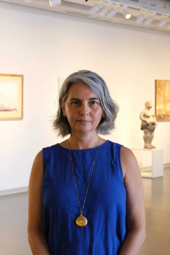 Ipek Cankaya, dalgalı gri saçları, kobalt mavisi kolsuz bluzu ve altın rengi uzun kolye ucuyla, arkasındaki aydınlık ve modern sanat galerisinde bulanık sanat eserleri ile heykellerin önünde kendinden emin bir şekilde duruyor.