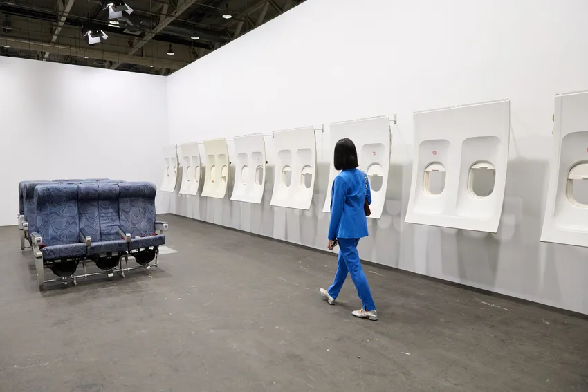 Beyaz bir galeride, mavi takım elbiseli, kısa saçlı bir kişi, solda düzenlenmiş mavi uçak koltukları ve duvara monte edilmiş bir dizi beyaz uçak pencere panellerinden oluşan Isa Genzken'in modern sanat eserine doğru yürüyor.