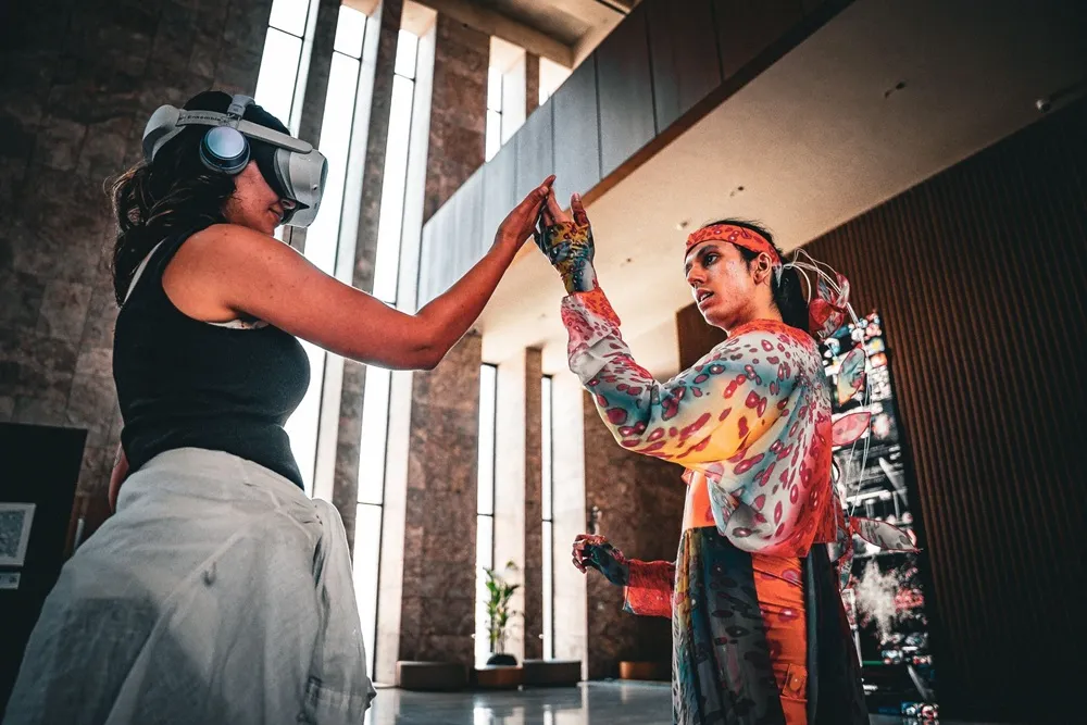 İstanbul Dijital Sanat Festivali'nde iki sanatçının etkileşimini gösteren bu fotoğraf, bir kişinin VR gözlük takarak dijital dünyaya adım attığını, diğerinin ise renkli giysiler ve el boyalarıyla fiziksel bir performans sergilediğini ve avuç içlerinin hafifçe birbirine değdiğini gözler önüne seriyor.