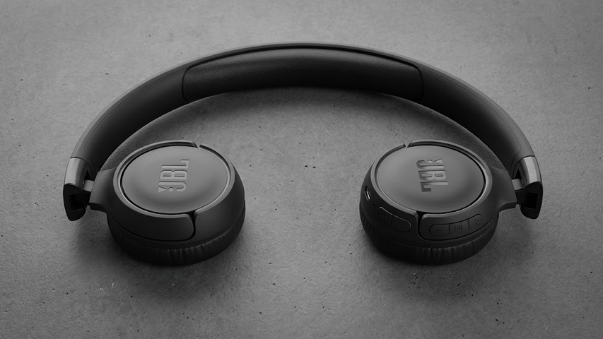 Koyu gri, hafif dokulu bir yüzey üzerinde, baş bandı zarifçe kavislendirilmiş, her iki kulaklığında da belirgin JBL logosu bulunan, siyah renkli JBL Tune 520BT kablosuz kulaklık yer alıyor; sağ kulaklıkta ses ayarı ve müzik kontrol düğmeleri net bir şekilde görülebiliyor.