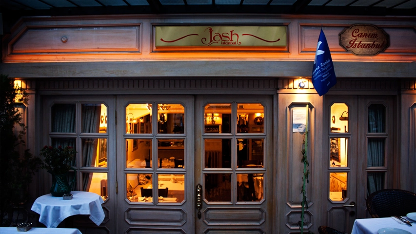 Kınalıada'da yer alan Jash İstanbul restoranının akşamüstü çekilmiş geniş bir görüntüsünde, ahşap cephesi, içeriden sızan sıcak ışıklarla aydınlanmış pencereleri ve dışarıdaki beyaz örtülü masaları ile Canım İstanbul yazılı tabelası belirginleşerek davetkar bir atmosfer sunuyor.