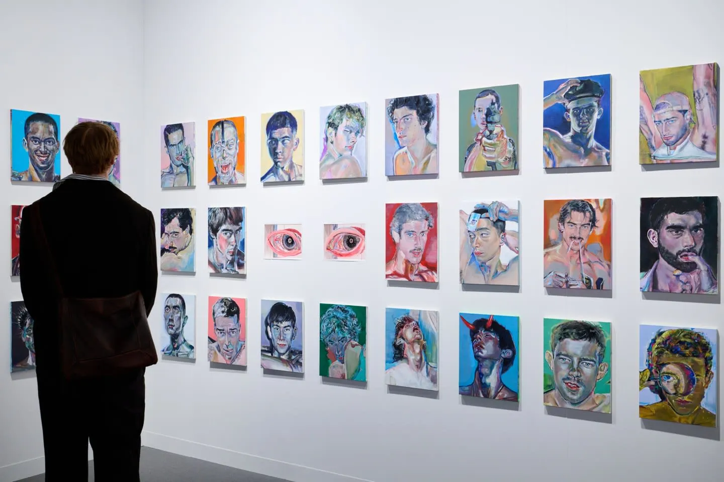 Bir sanat galerisinde sırtı dönük, koyu renk takım elbiseli ve çapraz askılı çantalı bir ziyaretçi, beyaz bir duvarda özenle sergilenen çok sayıda modern tabloya bakıyor. Sergi, Kosaku Kanechika'nın imzasını taşıyan, genellikle belirgin ifadeye sahip erkek portrelerini ve birkaç büyük göz resmini içeren, canlı renklerle yapılmış küçük ila orta boyutlu eserlerden oluşuyor, bazı portrelerde sürreal veya çarpıcı detaylar göze çarpıyor.