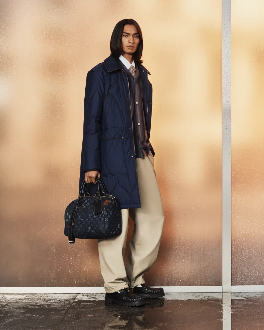 Genç bir erkek model, Louis Vuitton İlkbahar-Yaz 2027 görünümünü sergiliyor; lacivert kapitone uzun bir ceket, beyaz gömlek ve çizgili kazak kombinasyonuyla bej bol pantolon ve kalın tabanlı siyah mokasen ayakkabı giyen model, sağ elinde koyu mavi LV monogram desenli, kahverengi etiketli zarif bir çanta taşıyor.